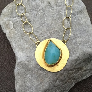 May include: A gold necklace with a large, teardrop-shaped turquoise stone set in a round, hammered gold pendant.