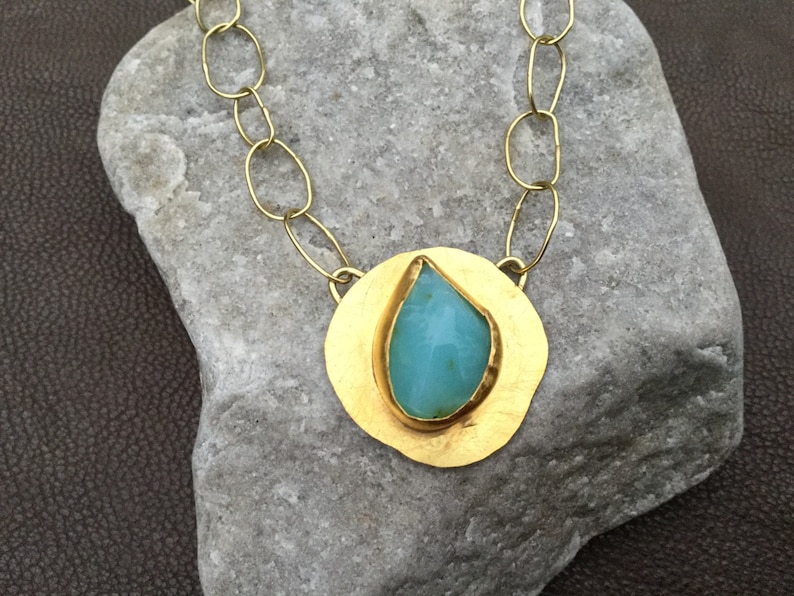 May include: A gold necklace with a large, teardrop-shaped turquoise stone set in a round, hammered gold pendant.