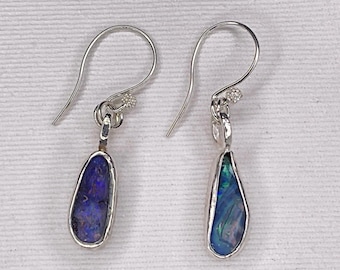 May include: A pair of silver drop earrings with teardrop-shaped, iridescent blue and purple stones. The stones are set in a silver bezel and hang from a silver hook with a small decorative element. The earrings are displayed on a white surface.