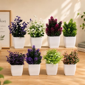 May include: A collection of eight small artificial plants in square white pots, arranged in two rows. The plants feature various foliage colors including deep purple, white, magenta, bright green, dark violet, and soft pink, set on a wooden surface.