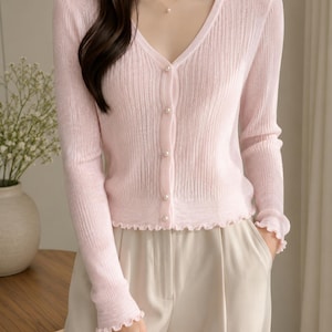 May include: A light pink, long-sleeved, ribbed knit cardigan featuring a V-neckline, a front button closure with small white pearl-like buttons, and delicate ruffled edges along the cuffs and the bottom hem of the garment.