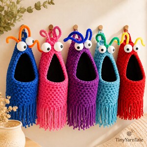 May include: Five hanging crochet wall organizers in blue, pink, purple, light blue, and red. Each features a wide open mouth, two large white googly eyes, and two curled antennae on top. The base of each organizer has a fringe of yarn tassels.