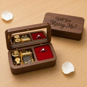Pode incluir: Uma caixa de música retangular de madeira escura com tampa articulada gravada com o texto Will You Marry Me? A caixa aberta revela um mecanismo musical dourado e um compartimento de veludo vermelho com um anel prateado com pedra clara.