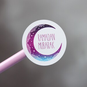 May include: A white round sticker with a purple and blue crescent moon design. The moon has stars and a white outline. The text "RAMADAN MUBARAK" is written in pink inside the moon.