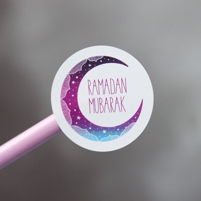 May include: A white round sticker with a purple and blue crescent moon design. The moon has stars and a white outline. The text "RAMADAN MUBARAK" is written in pink inside the moon.