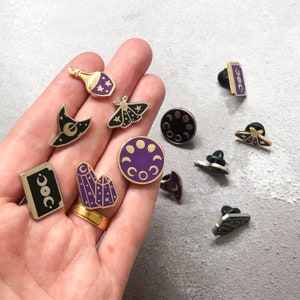 May include: A collection of 10 enamel pins featuring various celestial and witchy themes. The pins are gold-toned with purple accents and include designs such as a crescent moon, a witch's hat, a crystal, and a moth.