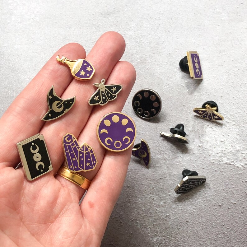May include: A collection of 10 enamel pins featuring various celestial and witchy themes. The pins are gold-toned with purple accents and include designs such as a crescent moon, a witch's hat, a crystal, and a moth.
