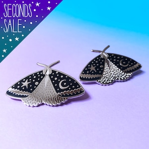 May include: Two silver-toned enamel pins shaped like moths with black wings and white accents. The moths have a crescent moon and a star on their wings.