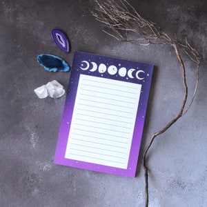 Peut inclure: Un bloc-notes violet et blanc avec un motif de phases lunaires et des pages lignées. Le bloc-notes est posé sur une surface grise avec deux tranches d'agate violette et bleue et un cristal de quartz transparent.