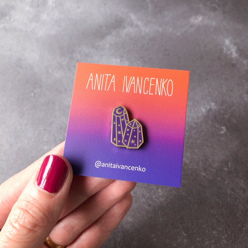 May include: Gold enamel pin featuring two purple crystal shapes with a crescent moon and stars inside. The pin is on a card with the text "Anita Ivancenko" and "@anitaivencenko".