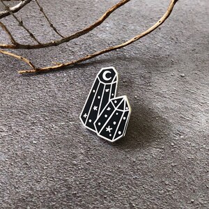 May include: Black enamel pin featuring two crystal shapes with a crescent moon and stars on a gray background.