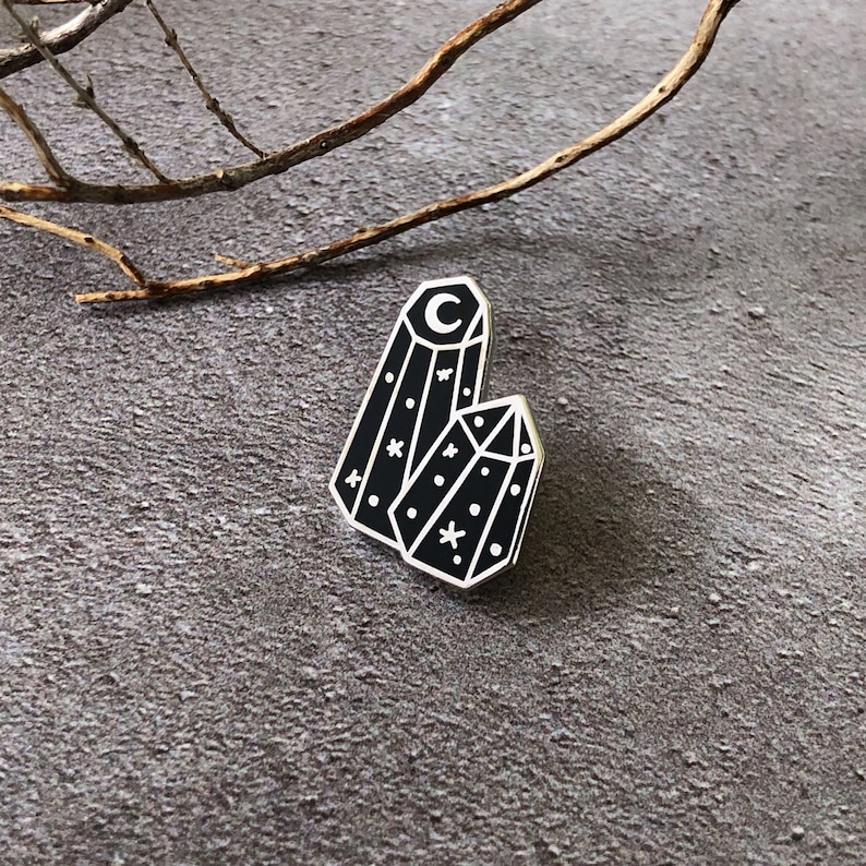 May include: Black enamel pin featuring two crystal shapes with a crescent moon and stars on a gray background.