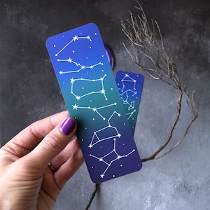May include: A blue and green gradient bookmark with white constellations printed on it. The bookmark is held in a hand.