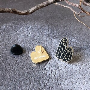 May include: Gold-tone enamel pin featuring a black crystal with a crescent moon and stars. The back of the pin is gold-tone with a heart shape.