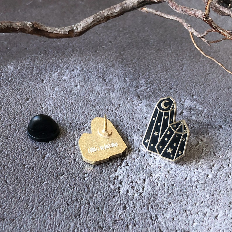 May include: Gold-tone enamel pin featuring a black crystal with a crescent moon and stars. The back of the pin is gold-tone with a heart shape.