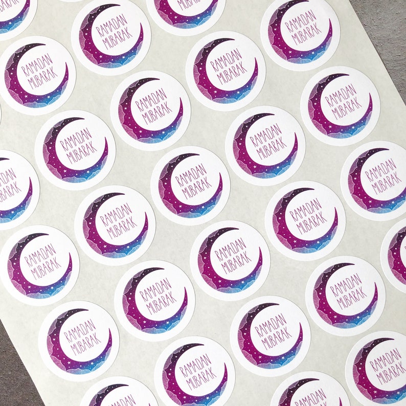 May include: A sheet of round stickers with a crescent moon design. The moon is purple and blue with white stars. The text "Ramadan Mubarak" is written in white on the moon.