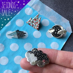 May include: Four enamel pins with different designs. The pins are silver with black and white details. The designs include a mountain, a cloud with a moon, a cloud with stars, and a geometric shape.