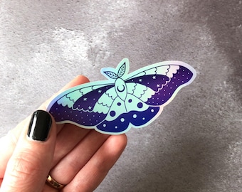 May include: A holographic sticker of a moth with a crescent moon on its head. The moth has a blue and purple gradient with white stars and dots.