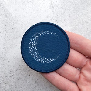 May include: A dark blue fabric patch featuring a white crescent moon design. The moon is filled with small white dots, resembling stars.