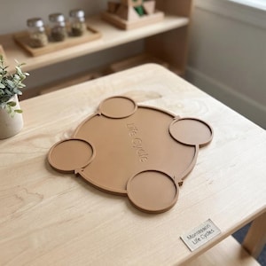 May include: A tan silicone life cycle mat featuring four circular sections connected by arrows in a circular flow pattern. The center of the mat is embossed with the text "Life Cycle" and a small rectangular label nearby reads "Montessori Life Cycles".