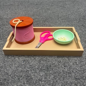 May include: A rectangular tan tray containing a pink spool of thread with a light yellow string, a pair of bright pink scissors with metal blades, and a small light green bowl holding several short pieces of light yellow string.