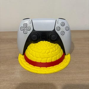 Op de afbeelding: Een wit en zwarte videogamecontroller die rust op een gele standaard in de vorm van een strooien hoed met een rode band. De controller is voorzien van standaard richtingsknoppen, twee zwarte analoge sticks en een PlayStation-logo.
