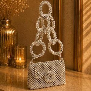 May include: A rectangular handbag covered in small, reflective silver beads. The bag features a flap closure with a circular beaded ornament at the center. The handle consists of a series of large, interlocking circular rings made of silver beads.