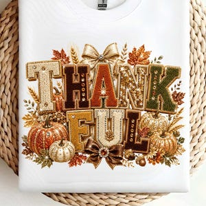 May include: A white crew neck t-shirt featuring a graphic print of the word THANKFUL in block letters. The letters are filled with a textured, sequin-like pattern in shades of gold, orange, and green, surrounded by illustrations of pumpkins and bows.