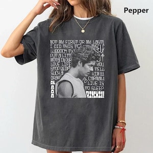 May include: A charcoal gray short-sleeve t-shirt featuring a white graphic print of a person in profile. The design includes text that reads: NOT MY FIRST OR MY LAST! I DID THIS TO MYSELF. LUBBOCK TX. PUT A LITTLE MOTA IN OUR LOVE LIFE. STOP BEING SUCH A DRAG. TELL HIM!!! CARNAL! LIVE IS TO SLEEP. RITCHIE!