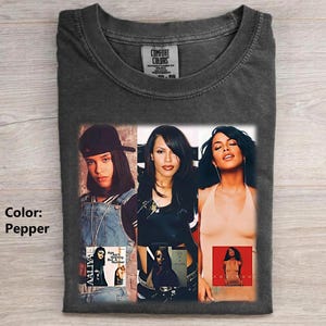 May include: A pepper-colored t-shirt featuring a graphic print of the singer Aaliyah in three different outfits. The design includes images of the artist wearing a baseball cap and denim overalls, a black top, and a beige halter top, with album art.
