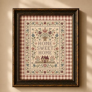 May include: A framed cross-stitch sampler featuring the text "HOME SWEET HOME" in black lettering. The design includes a central house, floral motifs, and hearts, all enclosed in a decorative border. The piece is set against a red and white gingham background and encased in a dark, distressed wooden frame.