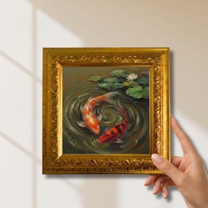 May include: A square oil painting in an ornate gold frame depicting two koi fish swimming in a circular motion within dark water. The scene includes green lily pads and a single white water lily floating on the surface of the pond.