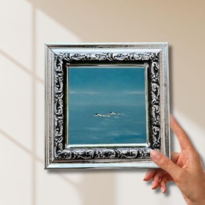 May include: A square painting in a silver-toned ornate frame with intricate scrollwork detailing. The artwork depicts a calm, expansive blue ocean scene with two small figures swimming in the distance under a clear, light blue sky horizon.