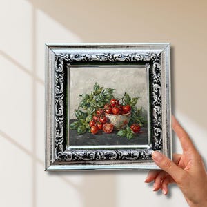 May include: An oil painting in a decorative silver frame depicting a bowl filled with red cherry tomatoes on the vine and green basil leaves. The artwork features a light gray textured background and a dark surface, capturing a still life scene.