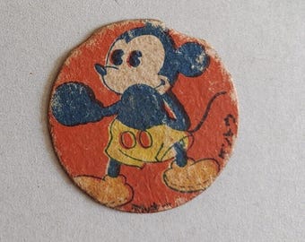 May include: A circular cardboard disc featuring a vintage illustration of Mickey Mouse in a boxing pose. The character is depicted in blue with yellow shorts, set against a textured orange background. The edges of the disc show signs of wear.