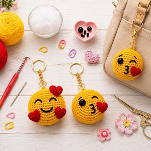 Può includere: Due portachiavi emoji rotondi all'uncinetto di colore giallo con cuori rossi, fissati ad anelli in metallo dorato. Un portachiavi raffigura un volto sorridente con due cuori, l'altro un volto che fa l'occhiolino con un cuore rosso.