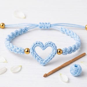 May include: A light blue macrame bracelet featuring a central crocheted heart motif, two polished gold-tone metal beads, and adjustable cord ends finished with gold-tone beads. The bracelet is accompanied by a wooden crochet hook and yarn.