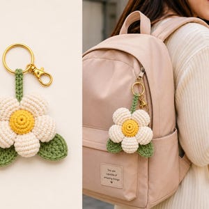 May include: A crochet flower keychain featuring five cream-colored petals, a bright yellow center, and two green leaves. The flower is attached to a green crocheted stem and a gold-toned metal split ring with a lobster clasp for easy attachment.