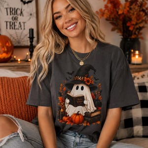 May include: A charcoal gray short-sleeve t-shirt featuring a graphic of a ghost wearing a witch hat with a leopard print band and sunflowers, reading a book titled Basic Witches, surrounded by orange pumpkins, autumn leaves, and bats.