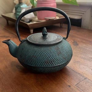 May include: A round cast iron teapot featuring a textured hobnail pattern in a dark teal and black finish. The teapot includes a matching circular lid with a small finial, a curved spout, and a sturdy, arched black metal handle attached to the top.