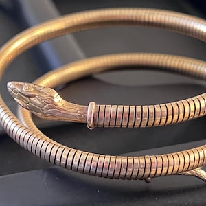 May include: A gold-toned metal wrap bracelet designed in the shape of a coiled snake. The bracelet features a textured, segmented body that mimics the appearance of scales, ending in a detailed, sculpted snake head with defined facial features.