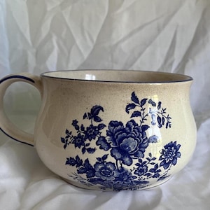 May include: A wide, cream-coloured ceramic mug featuring a speckled texture and a dark blue floral pattern. The mug has a rounded body, a single handle, and a dark blue rim. The design consists of detailed blue flowers and leaves.