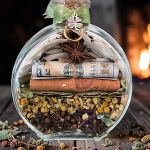 May include: A clear glass bottle with a cork stopper, tied with a green ribbon and a silver four-leaf clover charm. Inside are layers of dried chamomile flowers, cloves, bay leaves, a cinnamon stick, a rolled US dollar bill, and star anise.