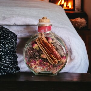 May include: A clear glass bottle with a cork stopper filled with dried rose petals, cinnamon sticks, and other natural ingredients. The bottle is sitting on a wooden surface.