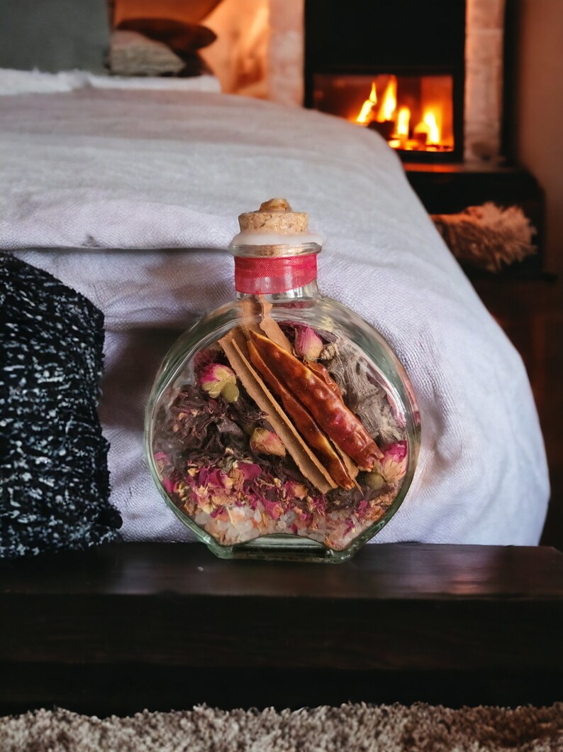 May include: A clear glass bottle with a cork stopper filled with dried rose petals, cinnamon sticks, and other natural ingredients. The bottle is sitting on a wooden surface.