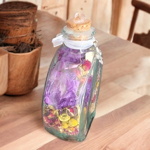 May include: A clear glass bottle filled with dried flowers in shades of purple, pink, and yellow. The bottle has a cork stopper and is tied with a white ribbon. A decorative charm hangs from the bottle.