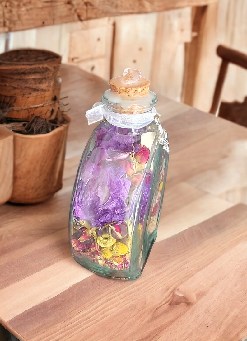May include: A clear glass bottle filled with dried flowers in shades of purple, pink, and yellow. The bottle has a cork stopper and is tied with a white ribbon. A decorative charm hangs from the bottle.