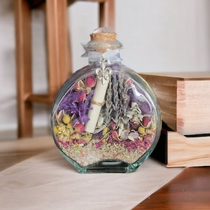 May include: A clear, round glass bottle filled with dried flowers, herbs, and a rolled-up scroll. The bottle has a cork stopper, a silver fleur-de-lis charm, and a light blue ribbon. The contents include lavender, rose petals, and other colorful botanicals.
