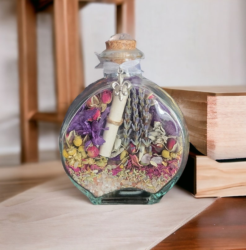 May include: A clear, round glass bottle filled with dried flowers, herbs, and a rolled-up scroll. The bottle has a cork stopper, a silver fleur-de-lis charm, and a light blue ribbon. The contents include lavender, rose petals, and other colorful botanicals.