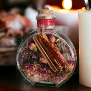 May include: A clear glass bottle with a cork stopper filled with dried herbs, spices, and rose petals. The bottle is sitting on a wooden surface.